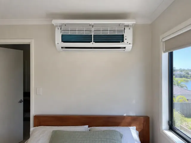 Clean split system indoor unit with open front panel above a bed in a bedroom with window and beige walls in Varsity Lakes